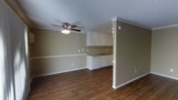 This single-floor space offers a seamlessly connected living and kitchen area designed for functional, everyday living. The open layout creates a sense of spaciousness, allowing natural sightlines to flow throughout the main living zone. Warm, dark hardwood-style flooring runs continuously across the space, providing a rich contrast against the neutral tan walls and crisp white trim and baseboards.

The kitchen is tucked neatly into a defined nook, framed by a decorative column with detailed molding. It features white upper cabinetry, a countertop with an integrated sink, and a dishwasher, all finished in a clean, classic style. A subtle under-cabinet light adds a practical touch to the workspace.

The living area is generously proportioned, with ample room for a variety of furniture arrangements. A ceiling fan with an integrated light fixture anchors the space, providing both airflow and ambient lighting. The textured popcorn ceiling and crown molding along the walls add an element of traditional character. Multiple electrical outlets and vents are thoughtfully distributed throughout, reflecting a well-planned layout. Overall, the space offers a comfortable and practical environment with a clean, move-in-ready aesthetic.
