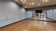 This dedicated fitness and dance studio is designed to accommodate a wide range of movement-based activities, from barre and dance classes to group fitness sessions. The space features warm-toned hardwood flooring throughout, providing a supportive and visually inviting surface ideal for active use. The walls are finished in a cool blue-gray palette, creating a calm and focused atmosphere conducive to exercise and instruction. A wall-mounted wooden ballet barre runs along one side of the room, offering a functional fixture for barre workouts and stretching routines. An expansive floor-to-ceiling mirror spans one full wall, maximizing the sense of space while allowing participants to monitor their form during classes. A large flat-panel display screen is mounted prominently above the mirror, enabling instructional content or visual guidance during sessions. Recessed and circular ceiling lights provide bright, even illumination throughout the studio. A bank of framed glass-panel doors connects the studio to the broader fitness facility, offering views into the main gym area and allowing for easy access and supervision. A wall-mounted thermostat and a security camera provide practical amenities, while the open, uncluttered layout ensures maximum usable floor space for participants of all disciplines.