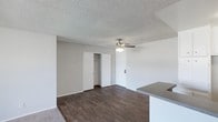 This single-floor space offers a clean, functional layout designed for comfortable everyday living. The main living area is finished with rich, dark hardwood-style flooring that adds warmth and contrast to the bright white walls and textured popcorn ceiling above. A ceiling fan with integrated lighting is centrally positioned, providing both air circulation and ambient light throughout the space.

A built-in closet with open shelving is conveniently located within the living area, offering practical storage directly accessible from the main room. Adjacent to this, a secondary door leads to additional areas of the unit.

The space transitions naturally into a kitchen area, partially visible through a raised counter bar with a grey laminate surface — a functional feature that separates the cooking and living zones while maintaining an open, connected feel. The neutral color palette throughout, consisting of white walls and trim paired with the dark flooring, creates a versatile backdrop that suits a variety of interior styles.

Overall, the space presents a straightforward and well-maintained interior, characterized by its cohesive finishes, practical storage solutions, and an open floor plan that maximizes the sense of space across the single level.