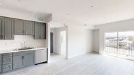 This single-floor unit presents a clean, contemporary design characterized by a bright, open layout and a cohesive neutral palette of whites and soft grays. Light wood-look flooring runs continuously throughout the space, reinforcing the sense of openness and flow between areas.

The kitchen is well-appointed with gray shaker-style cabinetry extending to the upper walls, offering generous storage. Stainless steel appliances — including a refrigerator, range, and dishwasher — complement the white countertops, creating a functional and modern workspace. Recessed lighting illuminates the area efficiently.

The living area is spacious and open, connecting seamlessly to a bedroom that features large sliding glass doors opening onto a private balcony. The balcony offers elevated views of the surrounding cityscape and distant hills, with ample natural light flooding the interior throughout the day.

Additional rooms are finished in soft gray tones, providing versatile spaces suitable for a variety of uses. The bathroom includes a shower with sliding glass doors, built-in open shelving, a gray vanity with a white stone-style countertop, and consistent gray tile flooring, maintaining the unit's cohesive modern aesthetic throughout.