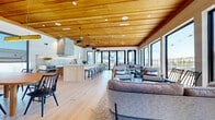 This contemporary single-floor community space showcases a thoughtfully designed open-concept layout that seamlessly integrates a full kitchen, dining area, and multiple lounge zones into one cohesive, flowing environment. The standout architectural feature is the striking warm-toned wood-plank ceiling with exposed beams, which adds natural texture and visual warmth throughout the entire space. Recessed lighting and pendant fixtures complement the ceiling, providing layered illumination across the interior.

The kitchen is equipped with light wood cabinetry, a large central island with bar seating, a prominent stone-clad range hood, and stainless steel appliances. Adjacent to the kitchen, a dedicated dining area features a solid wood table surrounded by sleek black chairs beneath large black-framed windows that bring in abundant natural light and views of the surrounding neighborhood.

The lounge areas are furnished with comfortable, contemporary seating arrangements, including generous sofas and accent chairs, all positioned to take full advantage of the floor-to-ceiling black-framed windows and sliding glass doors. These expansive openings frame direct views of an outdoor pool and patio area, flooding the interior with natural light. Light wide-plank hardwood flooring runs continuously throughout, tying all zones together in a clean, modern aesthetic. A wall-mounted television and built-in shelving round out the amenity offerings of this well-appointed space.
