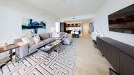This elegantly designed high-rise residence offers a seamless blend of modern architecture and refined interiors, all elevated by sweeping panoramic views of the city skyline and surrounding landscape. Light-toned hardwood flooring flows throughout the open-concept living and dining areas, anchored by a generous sectional sofa, a geometric-patterned area rug, and bold abstract artwork. Floor-to-ceiling windows and sliding glass doors lead to a private balcony, flooding the space with natural light.

The gourmet kitchen is appointed with dark espresso cabinetry, stainless steel appliances, crisp white countertops, and a central island with bar seating — a functional and stylish hub for entertaining.

The primary bedroom is a serene retreat, enveloped by curved floor-to-ceiling glass that frames breathtaking city views. A tufted headboard and plush bedding complement the soft, neutral palette. The ensuite bathroom features a freestanding soaking tub positioned beside panoramic windows, a glass-enclosed rainfall shower, and a double-sink vanity with wide mirror accents.

A secondary bedroom doubles as a versatile den or study, with corner windows offering additional city vistas. Throughout the residence, a consistent palette of white, grey, and dark wood tones creates a cohesive, sophisticated ambiance.