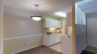This single-floor residence offers a practical and well-organized layout, with the kitchen serving as a central feature of the space. The kitchen is equipped with white cabinetry providing ample storage, a white dishwasher, a stovetop range, and a refrigerator. A double-basin sink is positioned beneath under-cabinet lighting, adding functionality to the workspace. The countertops complement the neutral color palette that runs throughout the space, which features warm beige and white tones on the walls and cabinetry. A fluorescent ceiling fixture illuminates the kitchen area, ensuring a well-lit environment for meal preparation. Adjacent to the kitchen, the dining or living area features dark carpeting and is lit by a stylish pendant light with a bowl-shaped shade, adding a subtle decorative touch. The transition between the tiled kitchen floor and the carpeted adjoining area creates a clear visual distinction between functional and living zones. The overall ambiance is understated and practical, with a clean, neutral aesthetic that provides a versatile backdrop for a variety of interior styles.
