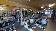 This fitness center offers a comprehensive workout facility designed to accommodate a variety of exercise routines. The space is outfitted with an extensive selection of cardio machines, including stationary bikes, ellipticals, and stair climbers, arranged to allow comfortable movement between equipment. A dedicated free weights area features a full rack of dumbbells and weight plates, catering to strength training needs. Large mirrors line the walls throughout the space, creating a sense of openness and allowing users to monitor their form during workouts. The flooring consists of durable rubber matting, providing both safety and comfort underfoot. Ceiling fans are installed throughout, promoting air circulation and maintaining a comfortable environment during intense workouts. Track lighting overhead ensures the entire floor is well-lit. The warm-toned walls contribute to an energizing yet welcoming atmosphere. The single-level layout ensures easy accessibility and seamless flow throughout the facility, making it a practical and functional space for fitness enthusiasts of all levels.