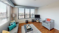 This 2-bedroom, 2-bathroom unit showcases a contemporary industrial-chic design highlighted by exposed silver ductwork, hardwood-style flooring in the main living areas, and large windows fitted with horizontal blinds that frame expansive urban views. The open-concept living area is bright and airy, benefiting from multiple oversized windows that flood the space with natural light while providing city skyline scenery.

The kitchen features dark espresso cabinetry extending to the ceiling, granite-style countertops, and a full suite of stainless steel appliances including a range, microwave, dishwasher, and refrigerator. Track lighting overhead ensures the kitchen is well-illuminated for cooking and entertaining.

Both bedrooms are versatile and well-proportioned, each equipped with ceiling fans, large windows with outdoor views, and built-in closets. The rooms accommodate loft-style or standard bed frames with under-bed storage, making efficient use of available space.

The two bathrooms mirror each other in design, each featuring dark vanity cabinetry, granite-topped sinks, large-format tile flooring, and tub-shower combinations. The neutral color palette throughout the unit creates a cohesive, modern atmosphere suited for comfortable everyday living.