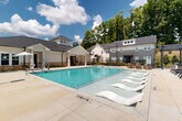 Located at 5740 Buffington Rd in Union City, Georgia, this outdoor amenity space centers around a generously sized, crystal-clear swimming pool that serves as the focal point of the community's recreational area. The pool features a gradual entry with in-water lounge chairs on one end, complemented by traditional pool access via stainless steel handrails on the other. Surrounding the pool is a broad concrete sundeck outfitted with multiple reclining lounge chairs, ideal for relaxing under the open sky. A covered pergola structure anchors one corner of the deck, providing a shaded gathering area furnished with Adirondack-style seating for a more casual, social atmosphere. On the opposite side, a white-exterior clubhouse building with a covered portico offers additional outdoor seating beneath market umbrellas, blending architectural charm with functional outdoor space. The community buildings feature a mix of white and dark gray siding with pitched rooflines, dormer windows, and brick accents, contributing to a cohesive and contemporary aesthetic. Mature trees line the perimeter, offering natural privacy and a lush green backdrop. The space is enclosed by black metal fencing, ensuring a secure and well-defined amenity area. The overall ambiance is bright, open, and inviting, reflecting a well-designed outdoor retreat.