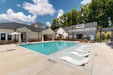 Located at 5740 Buffington Rd in Union City, Georgia, this outdoor amenity space centers around a generously sized, crystal-clear swimming pool that serves as the focal point of the community's recreational area. The pool features a gradual entry with in-water lounge chairs on one end, complemented by traditional pool access via stainless steel handrails on the other. Surrounding the pool is a broad concrete sundeck outfitted with multiple reclining lounge chairs, ideal for relaxing under the open sky. A covered pergola structure anchors one corner of the deck, providing a shaded gathering area furnished with Adirondack-style seating for a more casual, social atmosphere. On the opposite side, a white-exterior clubhouse building with a covered portico offers additional outdoor seating beneath market umbrellas, blending architectural charm with functional outdoor space. The community buildings feature a mix of white and dark gray siding with pitched rooflines, dormer windows, and brick accents, contributing to a cohesive and contemporary aesthetic. Mature trees line the perimeter, offering natural privacy and a lush green backdrop. The space is enclosed by black metal fencing, ensuring a secure and well-defined amenity area. The overall ambiance is bright, open, and inviting, reflecting a well-designed outdoor retreat.