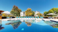 Located at 2002 Battlecreek Dr in Fort Collins, Colorado, this resort-style outdoor pool area offers a compelling blend of relaxation and recreation. The centerpiece is a generously sized swimming pool with crystal-clear water, complemented by a separate spa for additional comfort. The expansive concrete sundeck surrounds the pool and is lined with vibrant blue lounge chairs, providing ample space for sunbathing beneath Colorado's bright open skies. A handsome wood pergola anchors one end of the deck and shelters an outdoor grilling station, making it well-suited for social gatherings. Adjacent to the pergola, a shaded patio features dining-height tables and chairs beneath a large umbrella, ideal for outdoor meals or casual conversation. The pool building itself is clad in attractive natural stone veneer with large windows, adding an architectural warmth to the outdoor environment. Lush mature trees frame the entire area, bursting with rich autumn foliage that creates a scenic backdrop. The surrounding landscaping and manicured hedges lend a sense of privacy and seclusion. Together, these elements create a welcoming, amenity-rich outdoor retreat that is both functional and visually appealing.