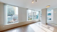 This contemporary single-floor residence is defined by its clean, modern aesthetic and abundant natural light. The open-concept living and dining area is anchored by an expansive wall of floor-to-ceiling windows, flooding the space with sunlight and offering sweeping urban views of the surrounding cityscape. Light hardwood flooring runs throughout the main living areas, complementing the bright white walls and adding warmth to the minimalist design. Track lighting overhead provides flexible illumination in the living space.

The kitchen is thoughtfully designed with a combination of warm wood lower cabinets and sleek white upper cabinetry, paired with a classic subway tile backsplash in soft blue-green tones. A full suite of stainless steel appliances — including a refrigerator, gas range, built-in microwave, and dishwasher — complete this functional and stylish culinary space.

A hallway lined with louvered storage doors connects the main living area to the private quarters, where the bedroom features plush carpeting and a sliding-door closet with built-in shelving. The bathroom is equipped with a full soaking tub and tiled surround with integrated shelving. Throughout, the unit maintains a cohesive, contemporary palette of neutral tones, creating a calm and inviting atmosphere.