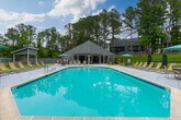 Located at 1001 N Donahue Dr in Auburn, Alabama, this outdoor amenity area offers a well-appointed community pool and patio space designed for relaxation and recreation. The centerpiece is a generously sized, diamond-shaped swimming pool with clear turquoise water, framed by a broad concrete deck that provides ample space for lounging. Rows of classic woven lounge chairs are arranged along the pool's perimeter, complemented by shaded seating areas with teal-colored market umbrellas, perfect for enjoying the outdoors in comfort.

A covered pergola structure anchors one side of the space, featuring exposed wood beams and an open-air design that offers shade while maintaining a connection to the surrounding landscape. Adjacent seating with cushioned chairs provides a relaxed gathering spot beneath the pergola. A life ring mounted to one of the support posts underscores the space's commitment to safety.

On the opposite side of the pool deck, a separate covered outdoor pavilion offers additional shaded seating with patio furniture, ideal for social gatherings. The entire area is enclosed by a fence and set against a backdrop of mature, towering trees that provide natural privacy and a serene, wooded ambiance. Well-maintained lawn areas and colorful landscaping complete the setting, creating an inviting outdoor retreat.