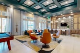 Located at 100 Oakbrook Drive in McDonough, Georgia, this single-floor community clubhouse is a vibrant and thoughtfully designed gathering space that blends contemporary aesthetics with comfortable functionality. The interior is anchored by a striking teal coffered ceiling that adds architectural depth and visual drama, complemented by large floor-to-ceiling windows dressed in sheer curtains that flood the space with natural light and offer views of the surrounding outdoor area.

Large-format porcelain tile flooring flows seamlessly throughout, providing a clean, polished foundation for the eclectic décor. The lounge area features bold retro-modern orange swivel chairs arranged around a sculptural wood coffee table, paired with a vibrant geometric area rug that adds warmth and definition to the space. A blue velvet sofa with colorful accent pillows adds an inviting social corner.

A dedicated billiards zone is set against a full-height wood-panel feature wall with integrated, backlit shelving, a wall-mounted flat-screen TV, and curated decorative art pieces. The bar area is a standout feature, with a curved white quartz countertop, warm wood cabinetry, artisan pendant lighting, and an eye-catching patterned tile backsplash. A wide glass-panel sliding door leads to an adjacent corridor, while modern wall art and a decorative console accent the entry. This space delivers a polished, resort-style atmosphere ideal for community gatherings.