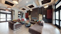 This single-floor space showcases a striking contemporary design characterized by an open, expansive layout and exceptional attention to detail. The ceiling is a standout architectural feature, displaying an elaborate coffered pattern with dark inset panels and geometric detailing, complemented by drum pendant lighting that adds warmth throughout.

The main lounge area is anchored by a large, deep-toned sectional sofa and matching ottoman, accented by vibrant throw pillows in bold reds, yellows, and teals. A low, rounded dark coffee table serves as a centerful centerpiece. Flanking the primary seating zone are vivid red accent chairs that introduce energetic contrast against the neutral palette.

A dramatic dark tile accent wall houses a sleek linear fireplace, framed by two sculptural columns of stacked raw logs — a distinctive organic element that balances the space's modern aesthetic. Nearby, bold red damask wall panels add rich texture and visual depth alongside dark credenzas styled with decorative objects and table lamps.

A quiet nook features a bistro-style table and woven chairs positioned before floor-to-ceiling windows dressed in elegant draping curtains, flooding the area with natural light and outward views. Light wood-toned flooring flows throughout, tying together the varied zones into a cohesive, sophisticated environment.