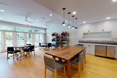 This well-appointed single-floor space is designed for versatile, communal use, seamlessly blending functional amenities with a warm, inviting atmosphere. The open-plan layout is anchored by warm-toned hardwood flooring that runs throughout, lending a cohesive, airy feel to the entire area.

The kitchenette is a standout feature, outfitted with crisp white shaker cabinetry accented by matte black hardware, gleaming white quartz countertops, and a striking arabesque-patterned tile backsplash. Stainless steel appliances, including a refrigerator and microwave, provide full functionality. Pendant globe lights hang above a substantial solid-wood communal dining table surrounded by woven chairs, creating an ideal gathering spot.

A more intimate seating area features a round table with upholstered navy blue chairs, a wall-mounted flat-screen television, and a sleek walnut credenza — well suited for small group meetings or casual viewing. Dedicated open shelving units display neatly organized yoga mats, resistance bands, and wellness accessories, indicating a dedicated activity or fitness-use zone.

Ceiling fans and recessed lighting maintain a comfortable, well-lit environment throughout. Large French doors and oversized windows flood the space with natural light and frame lush, verdant outdoor views, with direct access to an exterior patio — enhancing the overall sense of openness and connection to nature.