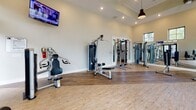 Located at 3340 Shipley St in Ladson, South Carolina, this fitness center is a comprehensive, single-floor facility designed to support a full range of workout routines. The space is defined by its impressively high ceilings and expansive floor-to-ceiling windows that line the walls, flooding the interior with abundant natural light and offering open views of the surrounding community and lush greenery.

The main workout area features a dedicated cardio zone with a row of treadmills and elliptical machines, each equipped with individual screens for entertainment during exercise. Full-length mirrors run along one wall, visually expanding the space and allowing users to monitor their form. Wall-mounted televisions provide additional viewing options throughout the facility.

A separate section of the gym houses a variety of strength and resistance training equipment, including cable machines, weight benches, and multi-function strength stations. Cycling and rowing machines are also available, catering to a diverse range of fitness preferences and skill levels.

The flooring throughout consists of durable wood-style planks, contributing to a clean, modern aesthetic. Recessed lighting and pendant fixtures complement the natural light to ensure consistent illumination across both areas. The overall design is open, airy, and well-organized, creating an inviting environment for focused, effective workouts.