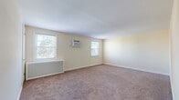 Located at 1 Garden Terrace in North Arlington, NJ, this single-floor unit offers a practical and well-organized layout across several rooms. The interior is finished with warm, neutral cream-toned walls and plush wall-to-wall carpeting throughout the living and bedroom areas, creating a cohesive and comfortable atmosphere.

The spacious main living area is notably generous, featuring multiple windows that invite abundant natural light and offer views of surrounding greenery. Each room is equipped with wall-mounted air conditioning units and decorative radiator covers, ensuring year-round climate comfort. The windows are fitted with blinds, providing privacy while still allowing light to filter through.

The kitchen is a functional standout, featuring warm honey-toned wood cabinetry, a gas range, a refrigerator, and a microwave — all in a coordinating black finish. Light hardwood-style vinyl flooring and sage green accent walls give the kitchen a welcoming, practical character, complemented by a window overlooking the outdoors.

The additional rooms are well-sized, each with natural light from windows and consistent neutral finishes. The clean white trim and baseboards throughout lend a polished, tidy appearance. Overall, the space presents a straightforward, comfortable residential layout suited for everyday living in the heart of North Arlington.