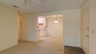 This single-floor residence offers a straightforward and functional layout designed for comfortable everyday living. The main living area is finished with wall-to-wall carpet and features a ceiling fan overhead, lending a casual and practical feel to the space. An open archway connects the living area to the dining space, creating a seamless flow between the two rooms. The dining area is highlighted by a classic pendant light fixture that hangs centrally, providing warm, focused illumination. Adjacent to the dining area, the kitchen is outfitted with white cabinetry and a matching countertop with a built-in sink, accented by a subtle tile backsplash that adds a touch of texture to the clean, neutral palette. The kitchen's white cabinetry and light-toned walls carry a cohesive, understated aesthetic throughout the home. In the main living area, a section of parquet wood flooring is visible, providing an interesting contrast to the carpeted zones and adding a layer of character to the interior. The overall ambiance of the space is calm and neutral, with a simple, open design that offers a solid foundation for personalization and decoration.