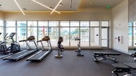 This contemporary fitness center is thoughtfully designed to support a full range of workout routines in a bright, open environment. The space is defined by its dramatic floor-to-ceiling window walls, which flood the interior with natural light and offer views of an outdoor pool area and surrounding landscape, creating an energizing atmosphere for exercise.

The cardio zone lines the windowed perimeter and is outfitted with multiple treadmills, stationary bikes, and elliptical machines, positioned to take advantage of the outdoor views. A dedicated free weights and functional training area features a comprehensive dumbbell rack, medicine balls, stability balls, and kettlebells, providing versatile options for strength and conditioning workouts.

The strength training section includes cable machines, weight benches, and additional resistance equipment arranged to maximize usable floor space. Mirrored walls in this zone enhance the sense of openness while supporting proper form during exercise.

Overhead, a distinctive cross-pattern linear lighting fixture adds a modern architectural accent to the high ceiling. Textured accent walls and warm-toned finishes contribute a polished, upscale aesthetic. The durable carpet flooring throughout is designed to withstand heavy equipment while providing comfort underfoot.