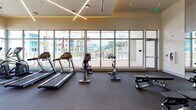This contemporary fitness center is thoughtfully designed to support a full range of workout routines in a bright, open environment. The space is defined by its dramatic floor-to-ceiling window walls, which flood the interior with natural light and offer views of an outdoor pool area and surrounding landscape, creating an energizing atmosphere for exercise.

The cardio zone lines the windowed perimeter and is outfitted with multiple treadmills, stationary bikes, and elliptical machines, positioned to take advantage of the outdoor views. A dedicated free weights and functional training area features a comprehensive dumbbell rack, medicine balls, stability balls, and kettlebells, providing versatile options for strength and conditioning workouts.

The strength training section includes cable machines, weight benches, and additional resistance equipment arranged to maximize usable floor space. Mirrored walls in this zone enhance the sense of openness while supporting proper form during exercise.

Overhead, a distinctive cross-pattern linear lighting fixture adds a modern architectural accent to the high ceiling. Textured accent walls and warm-toned finishes contribute a polished, upscale aesthetic. The durable carpet flooring throughout is designed to withstand heavy equipment while providing comfort underfoot.