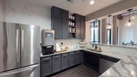 This single-floor space combines modern functionality with refined aesthetics, creating an inviting and well-designed environment. The kitchenette serves as a central highlight, showcasing rich charcoal-gray shaker-style cabinetry that extends from floor to ceiling, offering generous storage throughout. Stunning granite countertops with bold veining provide a striking contrast against the cabinetry, while a coordinating marble-look tile backsplash adds depth and elegance to the space.

Stainless steel appliances, including a full-size French door refrigerator and a built-in microwave, lend a professional quality to the kitchen area. A dedicated coffee and beverage station with a smart coffee machine makes this space ideal for communal or professional use. The undermount sink, paired with a matte black faucet, anchors a corner section that receives natural light through a nearby window framed by exposed brick accents and pendant lighting.

Dark wood-look tile flooring runs throughout, adding warmth and continuity to the space. The adjoining areas feature curated artwork — including botanical and abstract pieces — displayed against soft neutral walls with textured wallcovering accents. Globe pendant lights and recessed lighting provide layered illumination. The overall ambiance is polished yet welcoming, reflecting a thoughtfully designed communal or hospitality-oriented environment.