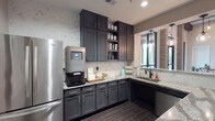 This single-floor space combines modern functionality with refined aesthetics, creating an inviting and well-designed environment. The kitchenette serves as a central highlight, showcasing rich charcoal-gray shaker-style cabinetry that extends from floor to ceiling, offering generous storage throughout. Stunning granite countertops with bold veining provide a striking contrast against the cabinetry, while a coordinating marble-look tile backsplash adds depth and elegance to the space.

Stainless steel appliances, including a full-size French door refrigerator and a built-in microwave, lend a professional quality to the kitchen area. A dedicated coffee and beverage station with a smart coffee machine makes this space ideal for communal or professional use. The undermount sink, paired with a matte black faucet, anchors a corner section that receives natural light through a nearby window framed by exposed brick accents and pendant lighting.

Dark wood-look tile flooring runs throughout, adding warmth and continuity to the space. The adjoining areas feature curated artwork — including botanical and abstract pieces — displayed against soft neutral walls with textured wallcovering accents. Globe pendant lights and recessed lighting provide layered illumination. The overall ambiance is polished yet welcoming, reflecting a thoughtfully designed communal or hospitality-oriented environment.