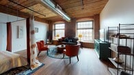 Located at 200 Esten Ave in Pawtucket, Rhode Island, this single-floor loft showcases the timeless appeal of industrial architecture beautifully blended with modern comfort. The open-concept layout flows generously from one area to the next, anchored by rich hardwood floors, dramatic exposed brick walls, and striking reclaimed wood beam ceilings that lend warmth and authenticity throughout.

The expansive living area is bathed in natural light streaming through large arched and multi-pane windows, framing views of the surrounding landscape. Exposed silver ductwork runs along the ceiling, reinforcing the space's industrial character. The living room is thoughtfully appointed with a mix of eclectic seating, including vibrant orange leather chairs and a neutral sofa centered around a patterned area rug.

The kitchen features a U-shaped layout with dark espresso cabinetry, granite countertops, and stainless steel appliances, offering both style and functionality. A secondary seating area with mid-century modern chairs and open shelving provides a versatile retreat within the loft.

The bathroom offers a refined contrast with its neutral geometric tile wainscoting, vessel sink vanity, and clean, spa-inspired finishes. Throughout the space, the interplay of raw industrial elements and curated décor creates a distinctive, inviting ambiance ideal for both living and entertaining.