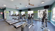 This fitness center offers a thoughtfully designed workout space that combines functionality with a clean, modern aesthetic. The room is finished with gray walls accented by a bold lime-green horizontal stripe, creating an energetic and motivating atmosphere. Large windows along multiple walls allow natural light to fill the space while providing views of the surrounding exterior.

The cardio section is prominently featured, housing a row of treadmills and elliptical trainers from Life Fitness, positioned to face wall-mounted televisions for entertainment during workouts. Rubberized flooring underlays the equipment, providing safety and sound absorption throughout the space. Additional strength and cross-training machines are positioned toward the perimeter, maximizing the use of available floor area.

Ceiling fans are installed throughout the room, ensuring consistent airflow and comfort during intense exercise sessions. A large mirror along one wall enhances the sense of openness and allows users to monitor their form. The overall layout is practical and well-organized, offering a range of equipment suitable for both cardio and strength training in a bright, inviting environment.