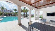 This inviting outdoor amenity space exemplifies resort-style living through its thoughtful architectural design and seamless connection to the surrounding tropical landscape. The covered pavilion is defined by a series of graceful white arched columns that frame panoramic views of a shimmering pool deck beyond, creating a elegant indoor-outdoor transition. Overhead, a rich wood-plank ceiling adds warmth and visual depth to the structure, complemented by recessed lighting for added ambiance. The light-toned tile flooring extends throughout the covered area, lending a clean and polished aesthetic. Outdoor dining is well accommodated with a generously sized table and chairs positioned beneath the pavilion, ideal for al fresco gatherings. A separate seating area with upholstered lounge furniture provides a comfortable retreat for relaxation. Beyond the columns, the pool area is lined with chaise lounges and surrounded by lush, mature palm trees that evoke a distinctly tropical atmosphere. The open sightlines allow natural light to flood the space throughout the day, while the surrounding landscape and water views create a consistently serene and resort-like ambiance. The architecture and finishes reflect a Mediterranean-influenced design sensibility, with crisp white stucco walls and arched detailing throughout.