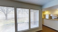This two-floor space offers a practical and comfortable layout designed to maximize usability across both levels. The living area is a standout feature, bathed in natural light through a wide bank of large windows fitted with white horizontal blinds, allowing for adjustable privacy and light control. The textured ceiling and neutral wall tones lend the room a clean, understated aesthetic, while the dark flooring provides a grounding contrast to the bright surroundings. Adjacent to the living area, a passthrough opening leads into the kitchen, which is finished with light-colored cabinetry and countertops. A range hood is visible overhead, and the warm yellow accent wall gives the kitchen a distinct, inviting character. The open sightlines between the living and kitchen areas create a connected, flowing feel throughout the main level. The overall ambiance of the space is light and airy, with an emphasis on natural illumination and functional design. Spanning two floors, the layout provides separation between living and private areas, making it a well-structured space suited for a variety of occupants and lifestyles.