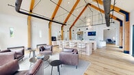 Located on Springside Lane in Middletown, CT, this expansive single-floor commercial space delivers an impressive blend of modern design and warm architectural character. The centerpiece is a dramatic great room anchored by soaring cathedral ceilings with exposed natural wood beams and contrasting black structural elements, creating a bold barn-inspired aesthetic. A large, light-toned island with bar seating serves as a functional hub, while globe and cone pendant lighting cascades from above. The great room flows into comfortable lounge seating areas with leather furnishings, a fireplace with marble surround, and floor-to-ceiling windows that flood the interior with natural light and offer outdoor views. An extended corridor lined with wide-plank light hardwood flooring connects multiple flexible seating zones, built-in shelving, and direct outdoor patio access through large glass doors. A welcoming reception area features a branded illuminated logo display and stylish lounge chairs. Private office spaces are equipped with granite-topped desks and floor-to-ceiling windows. The restrooms are finished with polished terrazzo floors, large-format wall tile, floating vanities, and sleek black hardware, completing this thoughtfully designed professional environment.
