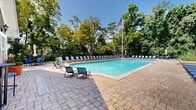 This two-floor property is defined by its impressive outdoor pool area and classic architectural character. The centerpiece of the exterior is a generously sized swimming pool with crystal-clear blue water, framed by a broad, paver-stone deck that provides ample space for relaxation and recreation. Rows of lounge chairs line the poolside, offering plenty of seating for sunbathing, while a pergola structure nearby provides a shaded retreat. A raised stone-wall terrace adds architectural interest and separates distinct zones within the outdoor space. The property is enveloped by mature, lush trees that create a sense of privacy and a tranquil natural backdrop, enhancing the resort-like ambiance. The main building showcases traditional shingle-style architecture with cedar shake siding, a steeply pitched roof, and multi-pane windows that bring abundant natural light into the interior. An adjacent pool house or auxiliary structure features large glass-paned doors and windows, connecting indoor and outdoor spaces seamlessly. An upper-level balcony with decorative iron railings extends from the main structure, overlooking the pool area. Together, the thoughtful landscaping, classic building design, and well-appointed outdoor amenities create an inviting and refined atmosphere throughout the property.