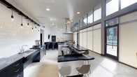 This striking single-floor space showcases a bold, contemporary aesthetic defined by clean lines and a sophisticated monochromatic palette. The open-plan layout centers around an expansive kitchen and dining area, where dramatic dark granite or stone countertops contrast sharply against crisp white walls and light polished concrete flooring. A large central island with built-in seating serves as the focal point, complemented by a sleek stainless steel range hood suspended overhead. Cabinetry and fixtures in deep charcoal tones reinforce the modern industrial character throughout. Overhead, an array of exposed pendant lights with bare bulbs adds a stylish, loft-inspired touch alongside recessed ceiling lighting. One of the most notable architectural features is the band of clerestory windows running along the upper portion of the wall, flooding the interior with abundant natural light while maintaining privacy. Roller shades offer flexible light control. A second seating zone with bar-height stools and a wall-mounted television creates a casual lounge area within the same open space. The overall ambiance is refined yet functional — a highly designed environment that balances dramatic visual impact with practical, well-appointed utility.