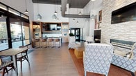 This contemporary single-level amenity space combines a café and social lounge into one cohesive, well-designed environment. The open layout is anchored by a café bar area, marked by illuminated "OPEN" and "CAFE" signage, featuring a counter with bar seating, wood-paneled cabinetry, a mounted flat-screen TV, and pendant lighting that adds a warm, industrial-chic character. Soaring ceilings with exposed ductwork contribute to the space's modern, loft-inspired aesthetic.

Adjacent to the café bar, a lounge area invites relaxation with stylish wingback chairs, patterned accent pillows, and a richly textured stone fireplace wall with a sleek linear gas fireplace. Built-in shelving displaying decorative books adds a library-like warmth to the nook, which also features upholstered bench seating, cylindrical concrete stools, and a wall-mounted TV for a versatile social setting.

A separate dining area bathed in natural light offers high-top tables and mixed seating alongside floor-to-ceiling windows and glass doors that open to street-level views. Framed artwork adorns the walls, reinforcing the space's curated, community-forward ambiance. The main entrance features double glass doors with a branded entry mat, welcoming visitors into this thoughtfully appointed space.
