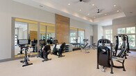Located on Sunset Blvd in West Columbia, SC, this fitness center offers a generously sized, single-floor space designed to support a comprehensive workout experience. The facility's architecture features notably high ceilings with recessed lighting and ceiling fans throughout, contributing to an open, airy atmosphere that feels expansive and comfortable during use.

The main exercise area is thoughtfully organized to accommodate a wide variety of equipment. A row of stationary and recumbent bikes lines one section, positioned in front of large floor-to-ceiling mirrors framed by warm wood accent panels, which enhance the sense of depth in the space. Strength training equipment, including cable machines, bench press stations, and free weight racks, is distributed throughout, providing ample room to move between stations.

A dedicated cardio zone features treadmills and an elliptical trainer positioned beneath oversized windows that flood the area with natural light and offer views of surrounding greenery. Stability balls and resistance accessories are neatly organized nearby. Additional multi-function strength machines occupy a separate section, maximizing the facility's training versatility. Light-toned flooring runs throughout, complementing the neutral wall palette and maintaining a clean, modern aesthetic.