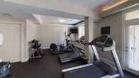 This fitness center is a comprehensively outfitted facility designed to support a full range of workout routines. The space is organized across a single floor and divided into distinct functional zones, allowing for an efficient and comfortable exercise experience.

The cardio area features an array of treadmills, elliptical trainers, and stationary bikes, each equipped with individual monitors for entertainment during workouts. Natural light filters in through windows fitted with blinds, complementing the flush-mounted ceiling lighting that keeps the space bright and well-illuminated throughout.

The strength training section is generously appointed, offering a cable machine system, a weight bench, and a dumbbell rack with a varied selection of free weights. Large wall-to-wall mirrors are strategically placed throughout the facility, visually expanding the space and allowing users to monitor their form during exercise.

A dedicated section with rubber flooring provides a safe, cushioned surface for floor exercises and functional training. An adjoining entryway area includes two private rooms — likely restrooms or changing facilities — alongside a community bulletin board displaying fitness schedules and notices. The neutral gray and white color palette, combined with dark rubber flooring, gives the center a clean, professional aesthetic that is both functional and inviting.