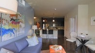 This single-floor space showcases an open-concept layout that seamlessly connects the living area to the kitchen, creating a cohesive and functional environment. The living area features a blue sofa accented with decorative pillows, anchored by an orange area rug and a coffee table styled with books and décor. Colorful wall art adds personality to the space. The kitchen is a standout feature, equipped with dark cabinetry that extends to the ceiling, stainless steel appliances including a refrigerator and range, and a tile backsplash. Pendant lights hang above a light-colored kitchen island with bar seating for three, providing both additional prep space and a casual dining option. Track lighting illuminates the kitchen workspace effectively. The polished concrete flooring runs throughout, lending an industrial-modern character to the space. The open layout allows sightlines from the living area straight through to the kitchen, enhancing the sense of spaciousness. A doorway visible on the right side leads to additional areas of the unit, while the overall dark-and-light contrast in the design choices creates a bold, contemporary ambiance.