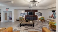 This single-floor amenity space is designed as a multifunctional social hub, seamlessly blending comfort and contemporary style. The main lounge area anchors the space with a striking exposed brick accent wall framing a wall-mounted flat-screen TV, flanked by curated black-and-white photography. Mid-century modern armchairs in olive green surround a rustic wood coffee table, all set atop a geometric patterned area rug. A leather sofa and pendant light fixture with an industrial cage design add character and warmth to the room.

The space opens into a dining zone featuring a solid wood communal table with upholstered chairs, ideal for gatherings. Adjacent to this, a billiards table offers additional recreational options, with views extending toward lush greenery through large windows that bring in abundant natural light.

The kitchen is a standout feature, boasting warm wood cabinetry paired with crisp white quartz countertops, including a generous central island. Globe pendant lights hang overhead while stainless steel appliances, a built-in wine rack, and open shelving complete the well-appointed setup. A mirrored backsplash element adds depth to the space. Large windows offer pleasant outdoor views, reinforcing the bright, airy atmosphere throughout this thoughtfully designed communal retreat.