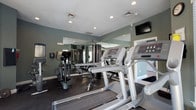 This fitness center is designed to support a comprehensive workout experience within a single-floor layout. The space is outfitted with a range of cardio equipment, including treadmills and elliptical machines, alongside a multi-station strength training unit, catering to a variety of exercise routines. Wall-to-wall mirrors line one side of the room, visually expanding the space and allowing users to monitor their form during workouts. A wall-mounted flat-screen television provides entertainment during exercise sessions. The flooring features a dark, durable surface well-suited for high-traffic fitness use. Recessed lighting in the textured ceiling ensures the room is well-lit throughout. The walls are painted in a muted sage green tone, contributing to a calm and focused atmosphere. Ventilation is provided through ceiling vents, maintaining air circulation during intense workouts. The overall layout is practical and efficient, with equipment arranged to allow comfortable movement between stations. This fitness center offers a functional and welcoming environment for a range of fitness levels and workout preferences.