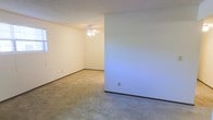 This single-floor residence offers a straightforward and functional layout designed for comfortable everyday living. The main living area is finished with wall-to-wall carpet in a neutral beige tone, providing a soft and warm underfoot feel throughout the space. Walls are painted in a clean, light neutral color that reflects available light and creates a bright, open atmosphere. Dark baseboard trim provides a subtle contrast against the lighter walls and flooring, adding a modest architectural detail that frames the room. A ceiling fan with an integrated light fixture is centrally mounted, offering both overhead illumination and air circulation. The layout includes an open adjoining area, suggesting a connected flow between living spaces. Electrical outlets are positioned at standard intervals along the walls, and a window allows for natural light to enter the space. The textured ceiling adds a classic residential touch. Overall, the space presents a clean, move-in-ready environment with a simple, no-frills design that provides a versatile canvas for a variety of interior arrangements and personal customization.