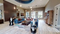 This single-story leasing office is designed to be both functional and welcoming, combining professional workspaces with comfortable communal areas. The main lounge serves as the centerpiece, featuring soaring ceilings with a distinctive circular tray detail finished in deep teal, complemented by an array of pendant lights that cast warm illumination across the space. Light wood-look tile flooring runs throughout, creating a cohesive flow between areas.

The lounge is furnished with teal sofas, accent chairs in rust and gray tones, and a rustic wooden coffee table anchored by a geometric area rug. Decorative open shelving units displaying curated artwork and objects line one wall, adding visual interest. Large windows and glass doors flood the space with natural light and offer direct views of an outdoor pool area.

A dedicated dining area features a round dark table with upholstered chairs beneath a circular ring pendant light, ideal for informal meetings. The private leasing offices are equipped with dark wood desks, ergonomic seating, and framed artwork. A kitchenette at the rear includes dark cabinetry, granite countertops, stainless steel appliances, and a tile backsplash, providing staff with a practical support area.
