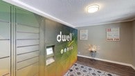 The Dwell Apartments lobby is a thoughtfully designed single-floor common area that combines practical amenities with a warm, inviting atmosphere. The centerpiece of the space is a large, eye-catching automated package locker system, wrapped in a bold, colorful geometric mural branded with the Dwell Apartments logo — making it both a functional and visually striking feature. The lockers are equipped with a touch-screen interface, offering residents a convenient and secure way to receive deliveries.

The main lobby area is finished with durable wood-style plank flooring and a textured ceiling, giving the space a clean, modern feel. A patterned area rug anchors the entryway, adding a layer of comfort and design detail. Natural light filters in through windows and a glass-paneled exterior door, brightening the interior and providing a view of the surrounding grounds.

A dedicated seating nook tucked alongside the locker wall offers a comfortable spot for residents, furnished with two modern green swivel chairs and a small accent table topped with a decorative lamp. A stylish industrial-style wall clock and motivational canvas wall art add personality and warmth to the space. A community bulletin board is also accessible near the entrance, ensuring residents stay informed. The overall ambiance is cheerful, functional, and welcoming.
