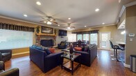 This single-floor clubhouse is designed to accommodate a variety of social gatherings, offering a generous, open layout with distinct functional zones that flow seamlessly into one another. The main lounge area serves as the centerpiece of the space, anchored by a striking stone fireplace with a wooden mantel and a wall-mounted flat-screen television. Multiple leather sofas and armchairs are arranged around glass-top coffee tables and area rugs, creating a comfortable and inviting seating arrangement suited for both casual relaxation and larger group events.

Hardwood flooring runs throughout the entire space, complemented by crown molding and dual ceiling fans with recessed lighting overhead. Natural light filters in through multiple windows dressed with decorative valances and blinds, while a set of glass doors provides direct access to an outdoor area.

Adjacent to the lounge, a dining area with a table and chairs offers additional seating, and a shuffleboard table adds a recreational element to the space. A compact but well-equipped kitchen features rich wood cabinetry, granite countertops, a mosaic tile backsplash, black appliances, pendant lighting, and a breakfast bar with bar seating — making it fully functional for hosting and catering needs.