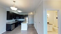 This single-floor space showcases a well-appointed kitchen that serves as its centerpiece. Dark espresso-toned cabinetry lines the walls, providing generous storage while creating a bold contrast against the light gray granite countertops. A central island with a white base and matching granite surface offers additional workspace and casual seating potential, topped by a stylish pendant light that anchors the space visually.

Stainless steel appliances, including a gas range, microwave, and refrigerator, are seamlessly integrated into the cabinetry, lending a sleek, contemporary feel. The wood-look plank flooring runs consistently throughout, adding warmth and cohesion. Crown molding along the ceiling perimeter adds a refined architectural detail, enhancing the overall sense of quality.

A wide hallway extends from the kitchen, lined with white panel doors — likely leading to storage closets or additional rooms — and features built-in cabinetry above, maximizing vertical storage. The neutral white walls and consistent lighting create a bright, clean ambiance throughout. The layout offers a natural flow between spaces, making the area feel open and functional.