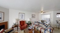 This two-story residence offers three bedrooms and two and a half bathrooms within a well-organized floor plan. The living room, pictured here, presents a bright and welcoming atmosphere, featuring neutral wall tones and light-colored carpeting that create a clean, airy backdrop. Vertical blinds on the windows allow for adjustable natural light throughout the day. A ceiling fan with an integrated light fixture provides both comfort and overhead illumination. The space comfortably accommodates a full seating arrangement, including a striped sofa and a classic wingback armchair in warm red tones, anchored by a wood coffee table. Framed artwork on the walls adds a finishing touch to the room's traditional aesthetic. The layout across two floors allows for a natural separation between living and sleeping areas, with the half bathroom conveniently located on the main level for guests. Overall, the residence combines practical design with a warm, traditionally styled interior, making it a functional and inviting living space.