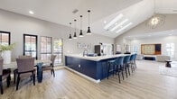 This single-story space is defined by its dramatic vaulted ceilings and open, flowing layout that connects several distinct functional areas under one roof. Light wood-look flooring runs continuously throughout, unifying the space and complementing the neutral gray walls.

The expansive kitchen is a standout feature, showcasing bold navy cabinetry with gold hardware, generous white quartz countertops, and stainless steel appliances including a large refrigerator and built-in microwave. A substantial navy island with seating for several provides an inviting bar-height gathering spot, illuminated by a cluster of matte black pendant lights. Skylights above the kitchen flood the area with natural daylight.

An adjacent dining area accommodates a large table and multiple chairs, framed by a row of black-framed windows offering exterior views. The living area features a shiplap accent wall, a wall-mounted television, and a mix of lounge seating including leather accent chairs and a sectional, all anchored by a patterned area rug.

A separate game lounge includes a large built-in Scrabble board, additional seating clusters, and decorative wall accents, making it ideal for social gatherings. Throughout, black-framed windows provide consistent natural light and a cohesive modern-farmhouse aesthetic.