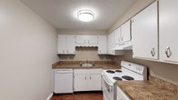 This single-floor unit offers a straightforward, functional layout across several well-defined living areas. The main living space greets you upon entry through a solid front door and is characterized by dark hardwood-style flooring, neutral taupe-toned walls, and white baseboards that create a clean, cohesive aesthetic. A window along the far wall allows natural light to filter in, brightening the open area. A wall-unit air conditioner provides climate control in this space.

The kitchen is efficiently designed in a U-shaped configuration, featuring white cabinetry both above and below granite-toned countertops. White appliances — including an electric range, dishwasher, and refrigerator — are neatly integrated, and a ceiling-mounted light fixture ensures the space is well-lit. Dark hardwood-style flooring continues throughout the kitchen.

The bedrooms offer a softer feel underfoot with plush neutral carpeting and are finished with the same warm taupe wall tones. Each room benefits from natural light through blinds-fitted windows.

The bathroom includes a full tub and shower combo with a white surround, a toilet, and dark hardwood-style flooring, keeping the space simple and easy to maintain. Overall, the unit presents a clean, move-in ready environment with consistent finishes throughout.