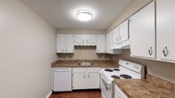 This single-floor unit offers a straightforward, functional layout across several well-defined living areas. The main living space greets you upon entry through a solid front door and is characterized by dark hardwood-style flooring, neutral taupe-toned walls, and white baseboards that create a clean, cohesive aesthetic. A window along the far wall allows natural light to filter in, brightening the open area. A wall-unit air conditioner provides climate control in this space.

The kitchen is efficiently designed in a U-shaped configuration, featuring white cabinetry both above and below granite-toned countertops. White appliances — including an electric range, dishwasher, and refrigerator — are neatly integrated, and a ceiling-mounted light fixture ensures the space is well-lit. Dark hardwood-style flooring continues throughout the kitchen.

The bedrooms offer a softer feel underfoot with plush neutral carpeting and are finished with the same warm taupe wall tones. Each room benefits from natural light through blinds-fitted windows.

The bathroom includes a full tub and shower combo with a white surround, a toilet, and dark hardwood-style flooring, keeping the space simple and easy to maintain. Overall, the unit presents a clean, move-in ready environment with consistent finishes throughout.