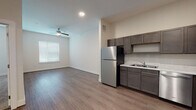 This single-floor unit offers a clean, modern design throughout its well-organized layout. The kitchen is a functional highlight, featuring dark gray cabinetry with brushed nickel hardware, light quartz-style countertops, and a full suite of stainless steel appliances including a refrigerator, electric range, microwave, and dishwasher. Recessed lighting illuminates the space, which transitions seamlessly into an open living area finished with wood-look plank flooring. A ceiling fan with a light fixture overhead and a window fitted with horizontal blinds provide comfort and natural light to the living space.

The bedroom offers a quieter aesthetic, with plush wall-to-wall carpeting, a ceiling fan, and a window that allows natural light to filter in. An additional room with wood-look flooring and a ceiling fan provides flexible use as a second bedroom or office.

The bathroom is finished consistently with the rest of the unit, featuring wood-look flooring, a dark-toned vanity with a single sink and light countertop, a standard tub and shower combination with built-in shelving, and neutral white walls. Throughout the unit, tall baseboards and a neutral color palette contribute to a cohesive, contemporary aesthetic.