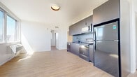 This well-designed 3-bedroom, 1-bathroom single-floor unit offers a clean, modern aesthetic throughout. Light wood-tone flooring runs continuously across the entire space, creating a cohesive and airy feel. The open-concept kitchen and living area serves as the heart of the unit, featuring a sleek, contemporary kitchen outfitted with dark matte cabinetry, stainless steel appliances including a refrigerator, electric range, microwave, and dishwasher, complemented by a stainless steel sink and backsplash. The living area receives generous natural light through large windows that frame expansive city and skyline views. A central hallway connects the three bedrooms, each finished with the same warm wood-tone flooring and crisp white walls. The primary bedroom is notably bright, boasting two large windows offering sweeping urban views and ample room to furnish. The additional bedrooms are well-proportioned, with one featuring a closet with a hanging rod for convenient storage. The shared full bathroom is finished with large-format tile flooring, white walls, a soaking tub with shower, and matte black hardware accents, lending a contemporary contrast to the otherwise light palette. The overall ambiance is clean, bright, and modern, well-suited for comfortable everyday living.