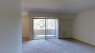 This single-floor residence offers a clean and functional layout designed for comfortable everyday living. The living area serves as the focal point of the space, finished with wall-to-wall carpeting that adds warmth underfoot. A large sliding glass door dominates one wall, fitted with vertical blinds that allow for adjustable light control throughout the day. The door opens directly onto a private balcony, where mature trees and greenery can be seen just beyond the railing, bringing a natural, tranquil element to the interior. Natural light filters generously into the living area through this expansive glass opening, giving the room an open and airy feel. The walls are finished in a neutral beige tone, providing a versatile backdrop suited to a variety of interior styles. A built-in wall vent is positioned near the sliding door, suggesting integrated climate control for year-round comfort. The overall design is understated and practical, with smooth ceilings and simple architectural lines that create a calm, uncluttered atmosphere. The space presents a solid foundation for personalization, with its neutral palette and connection to the outdoors being among its most notable features.
