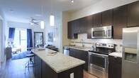 This single-floor space showcases a thoughtfully designed open-concept layout that seamlessly connects the kitchen and living area. The kitchen is a standout feature, equipped with rich dark cabinetry that extends to the ceiling, providing generous storage. A large granite-topped island anchors the kitchen, offering additional prep space and casual seating. Stainless steel appliances, including a built-in microwave, electric range, and dishwasher, complement the sleek aesthetic. A classic white subway tile backsplash adds a clean, timeless contrast to the dark cabinetry. Pendant lighting fixtures hang above the island, enhancing both function and style, while a ceiling fan in the adjoining living area promotes airflow throughout the open space. The living area extends naturally from the kitchen, creating a cohesive and practical flow ideal for everyday living and entertaining. Light-toned walls reflect ambient light, contributing to a bright and airy atmosphere. The combination of dark wood tones, stone surfaces, and stainless accents gives the space a contemporary, polished character throughout.